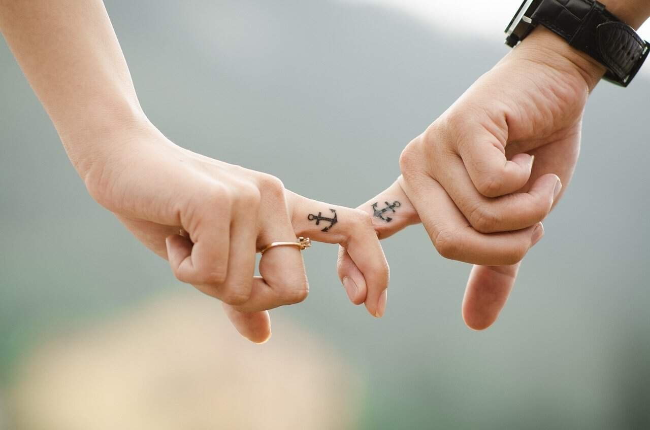 zodiac incompatibility Close-up photograph of two people linking pinky fingers against a soft, blurred background. One person wears a black watch, and both have small matching anchor tattoos on their fingers. One hand also wears a gold ring. The intimate gesture symbolizes connection and promises, ironically contrasting with the article's theme about zodiac incompatibility.