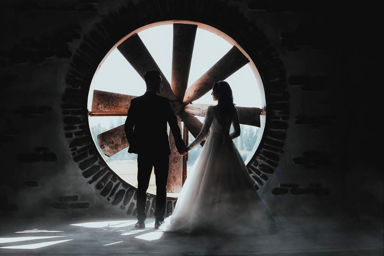 power couple zodiac Dramatic silhouette of a bride and groom standing before a large circular window with sunburst pattern. The couple is captured from behind, with the bride in a flowing white dress and groom in a dark suit. The majestic circular window bathes them in bright light against a dark stone surround, symbolizing the powerful union of two individuals. The striking composition and celestial-like circular frame suggests cosmic destiny and the astrological harmony of power couples brought together by the stars.